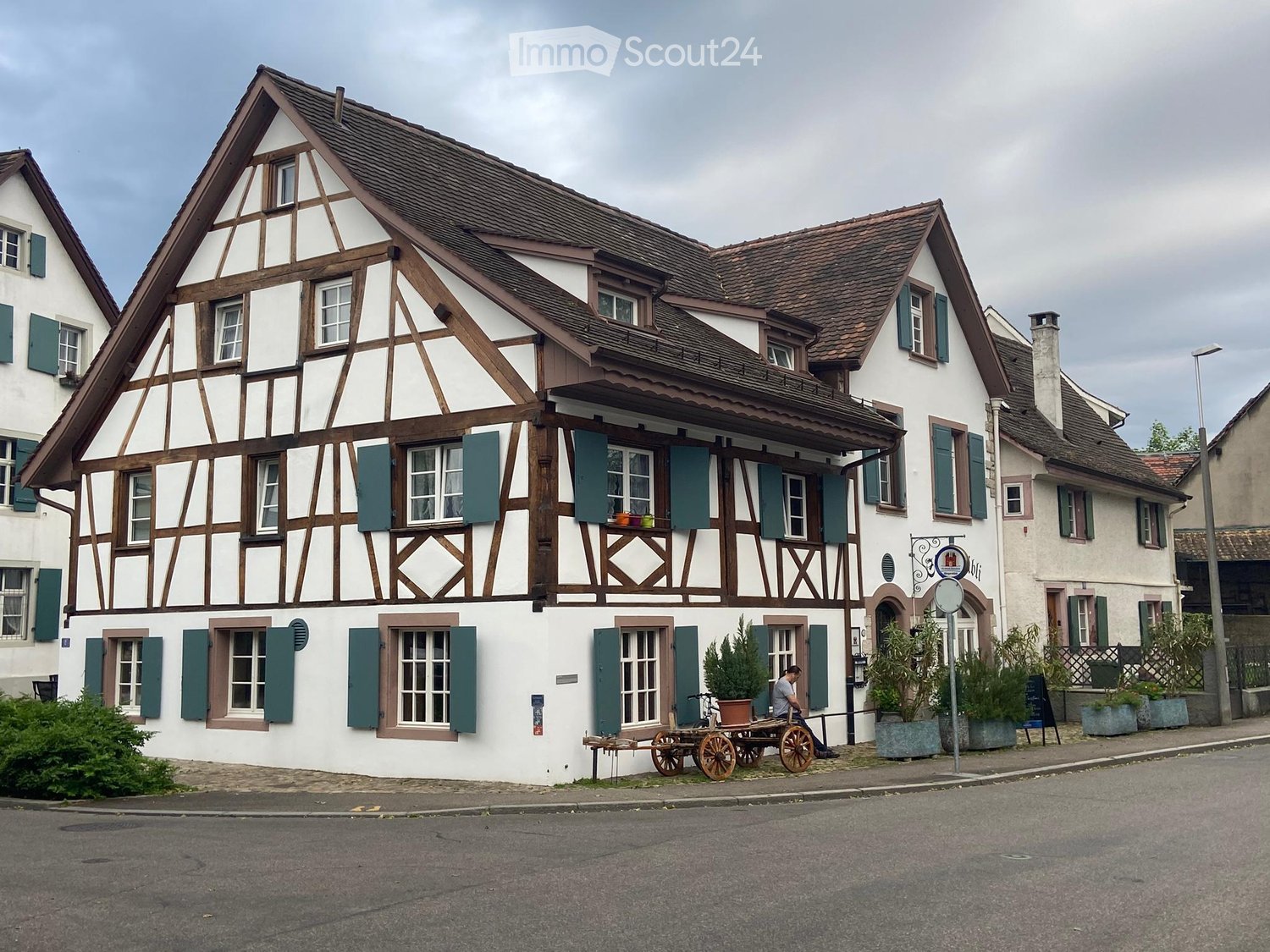 Traditional timber-framed house, brown roof, white walls, green shutters, brown trim, multiple windows, plants in front, man sitting on a cart