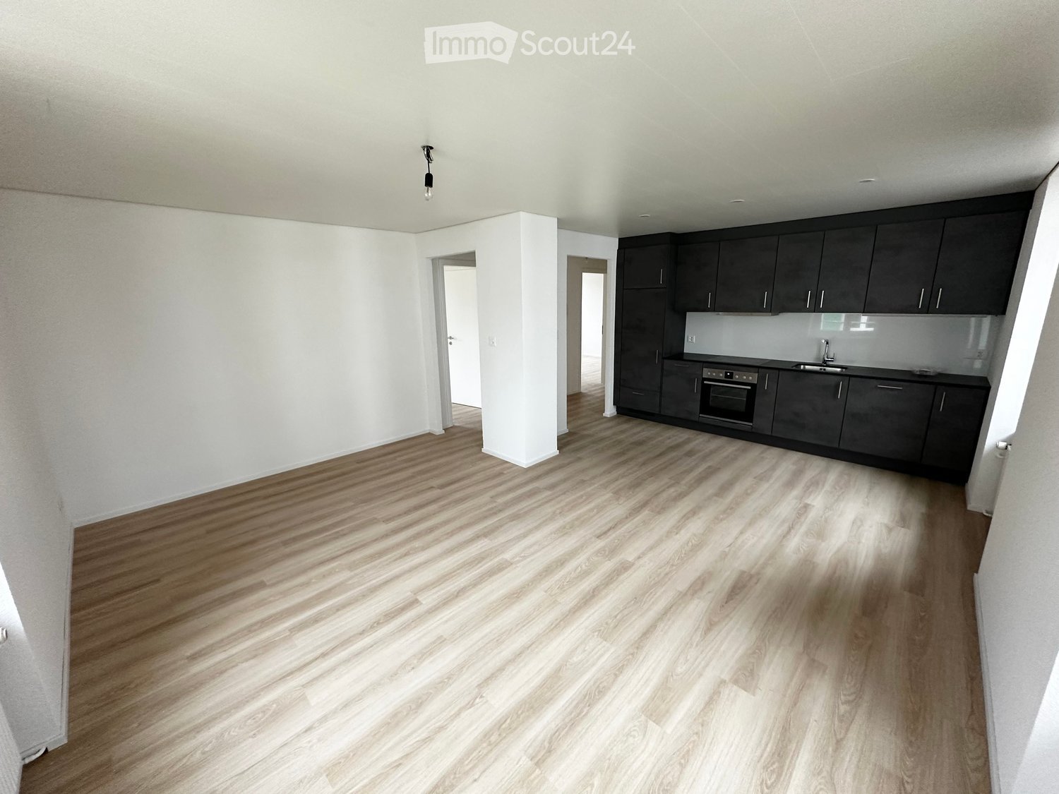 Empty room with light wooden flooring, white walls, and a small kitchen area with dark cabinets and appliances visible in the background.
