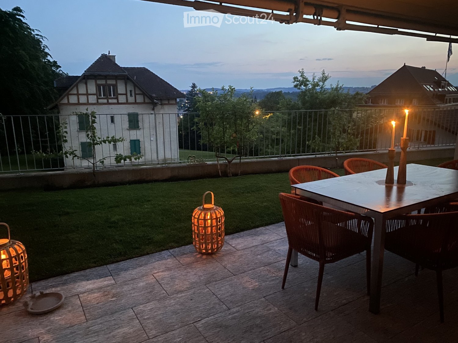 Balcony with wooden floor, metal railing, table and chairs, decorative lanterns, and a view of a house and landscape.