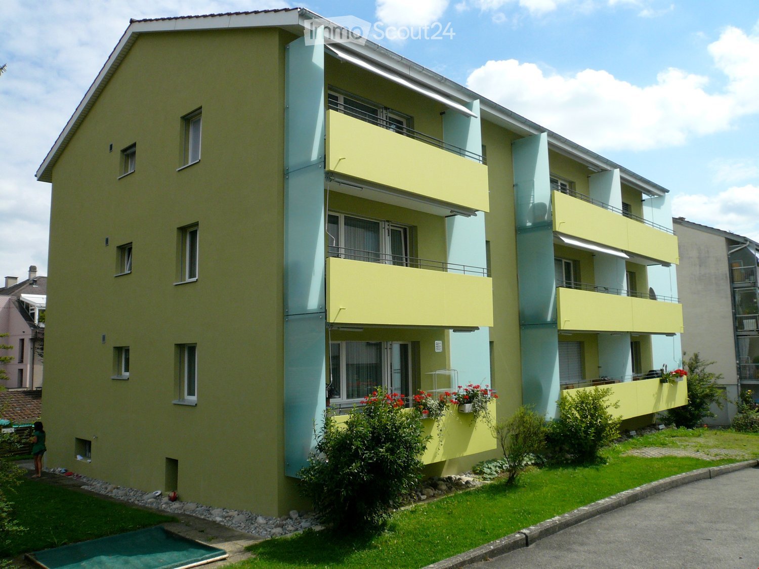A modern, two-story apartment building with yellow and light blue exterior walls, multiple balconies, and a small garden area with greenery and flowers.
