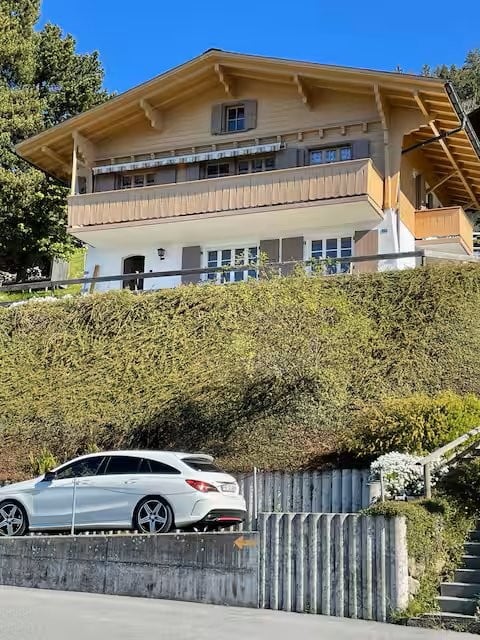 Two-story wooden house with balconies, surrounded by greenery, with a parked car in front.
