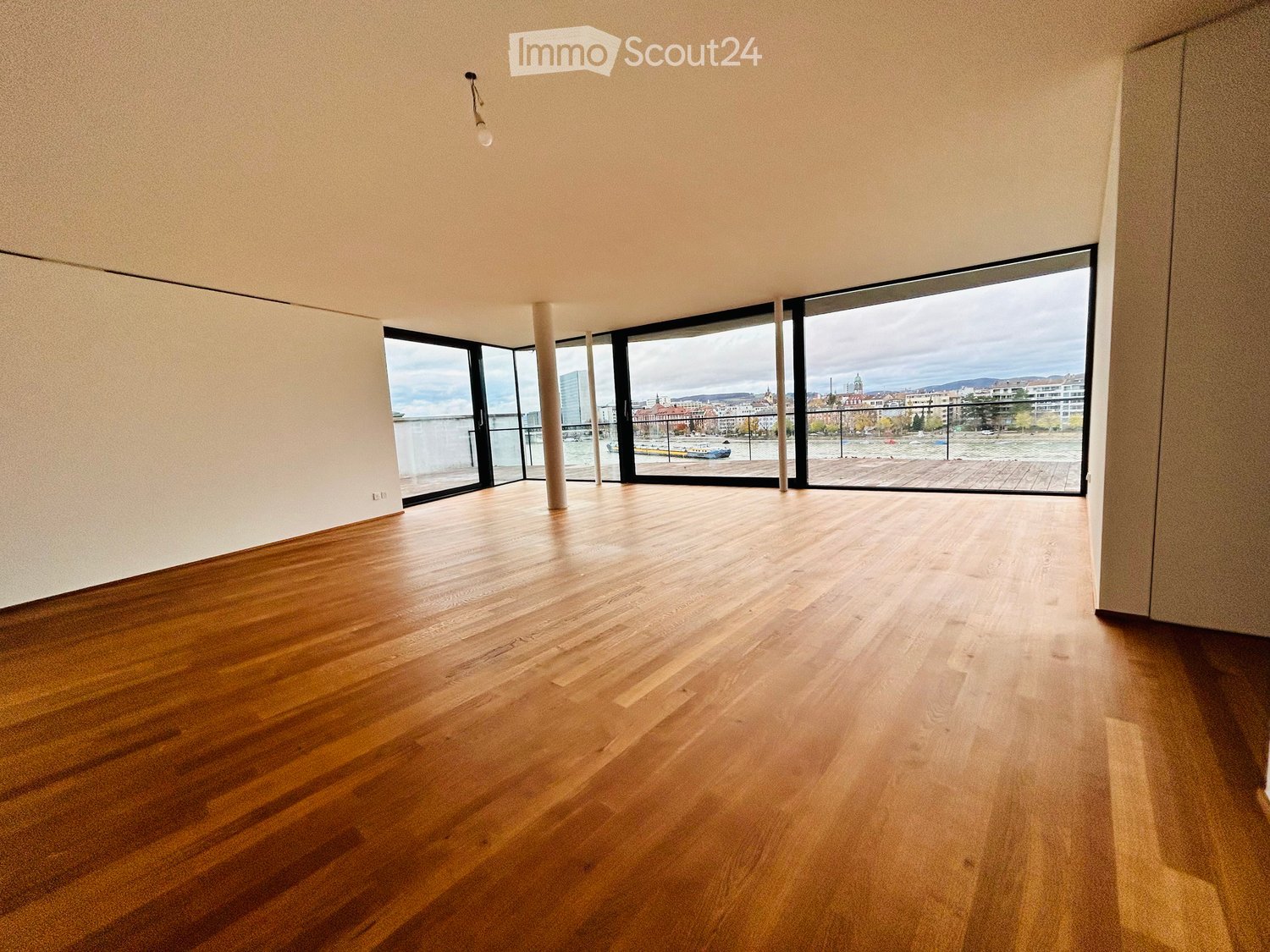 Empty room with wooden flooring, large windows, and a view of a river and cityscape.