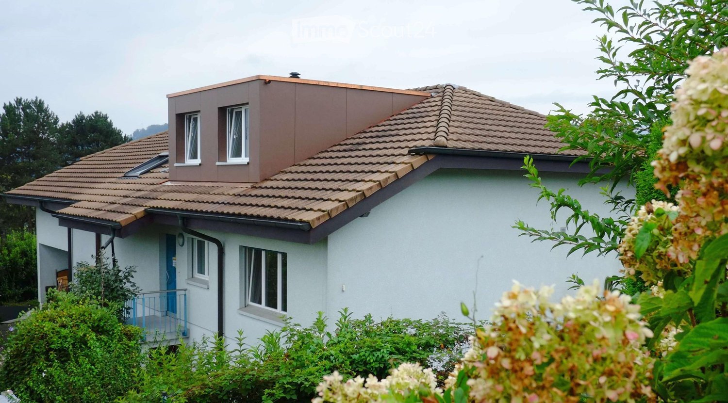 Two-story house with a tiled roof, white exterior walls, and multiple windows. Features a small balcony with a blue railing and a small garden area with greenery and flowers in the foreground.