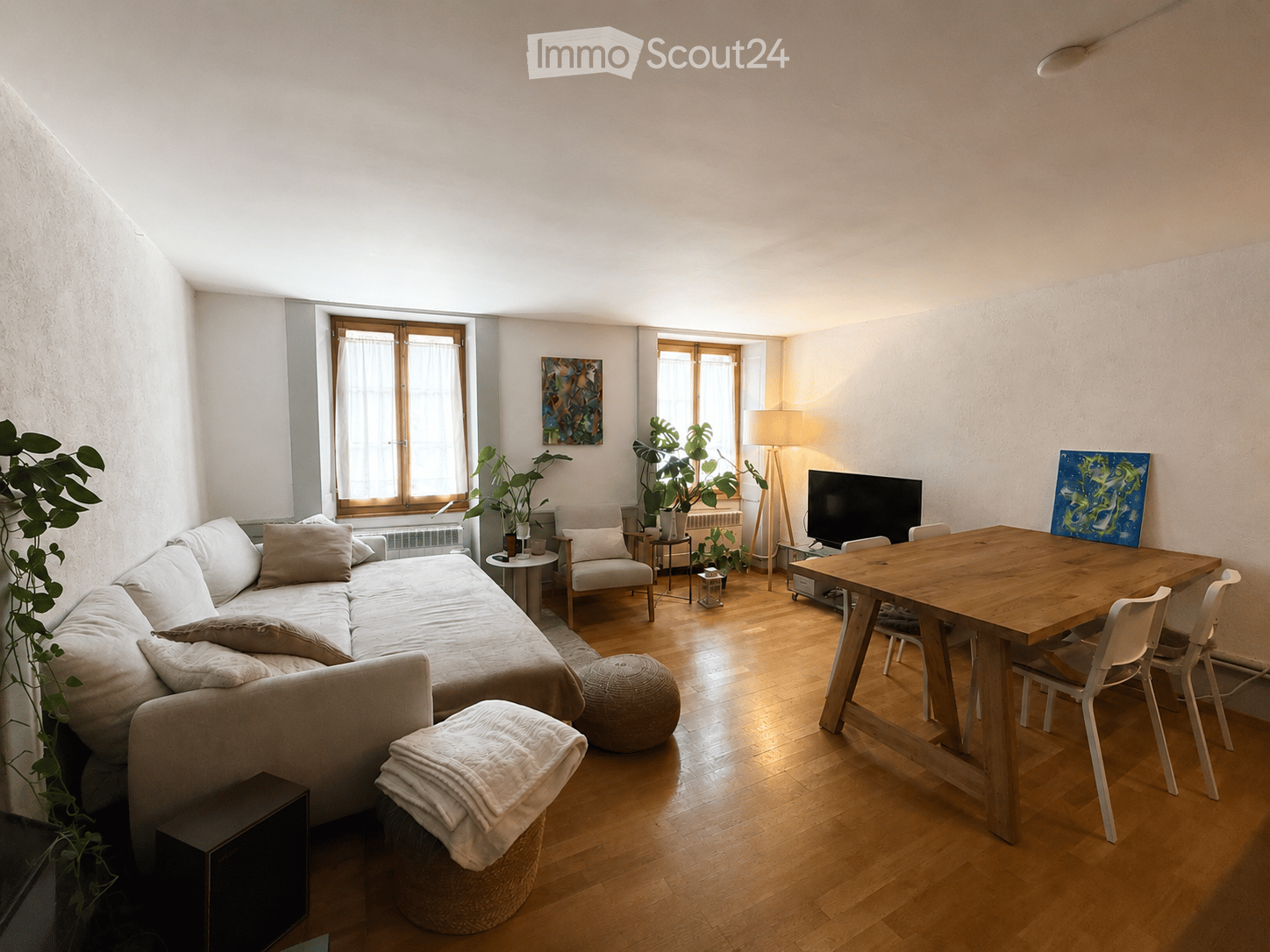 A modern living room with a sofa, coffee table, armchairs, a round wooden table with chairs, a floor lamp, and a television. The room features wooden flooring, large windows, and decorative plants.