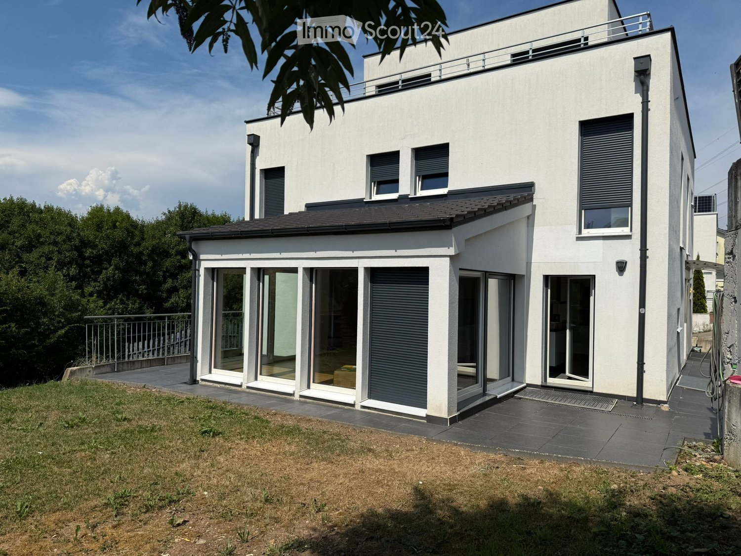 Modern two-story house with white exterior, large windows, and a raised ground floor. Features include a balcony on the upper level and a spacious patio area with glass doors leading to the interior.