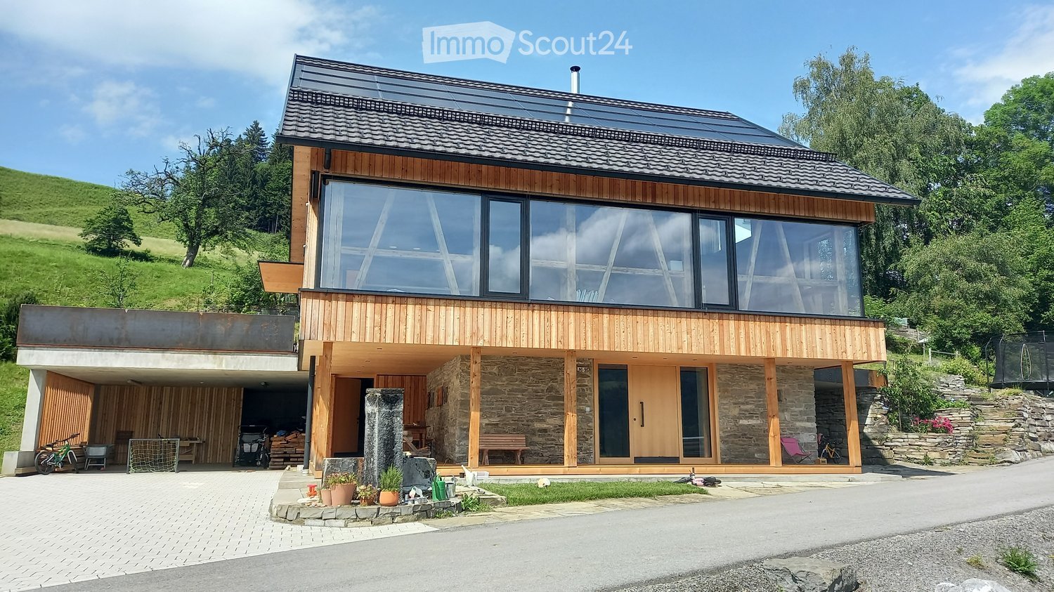 Modern two-story house with wooden and stone exterior, large windows, raised ground floor, garage, and outdoor seating area.