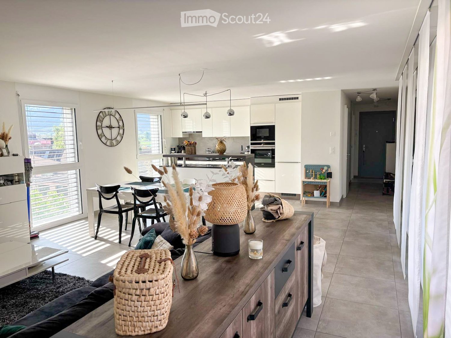 Open-plan living and dining area with modern furniture, large windows, and a kitchen area in the background.