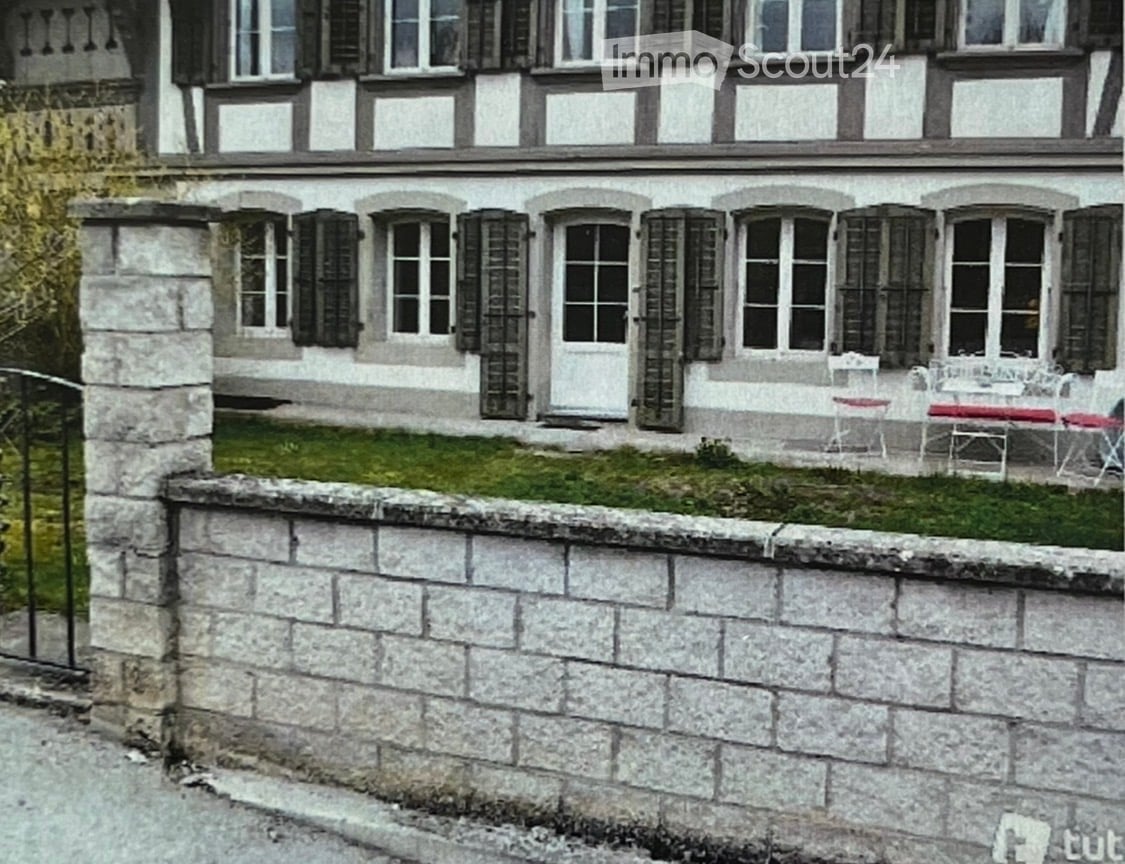Two-story building with white walls, dark shutters, and a raised ground floor accessed by steps. A small garden area with a table and chairs is visible in front.