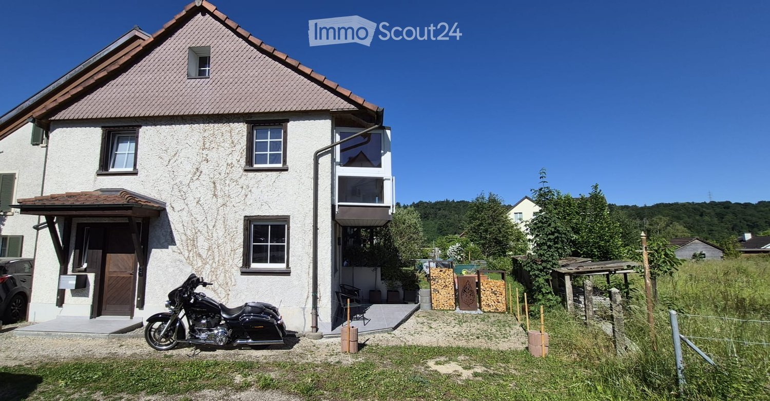Two-story house with white exterior, multiple windows, and a balcony on the upper floor. A motorcycle is parked in front, and there is a small garden area with a wooden structure and stacked firewood.