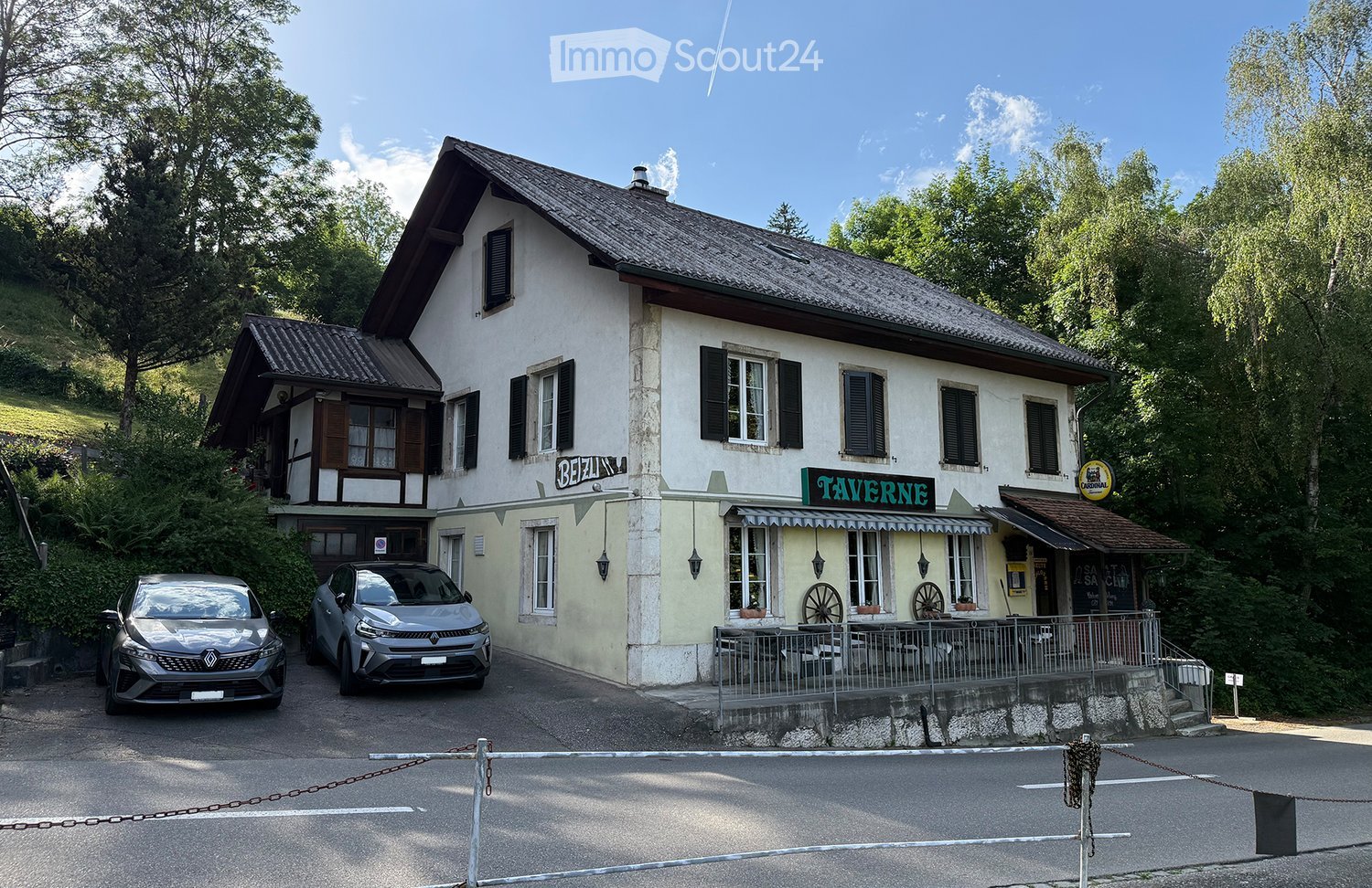 Two-story building with a sign reading "TAVERNE" and "BEIZLI" on the facade, surrounded by greenery and parked cars.