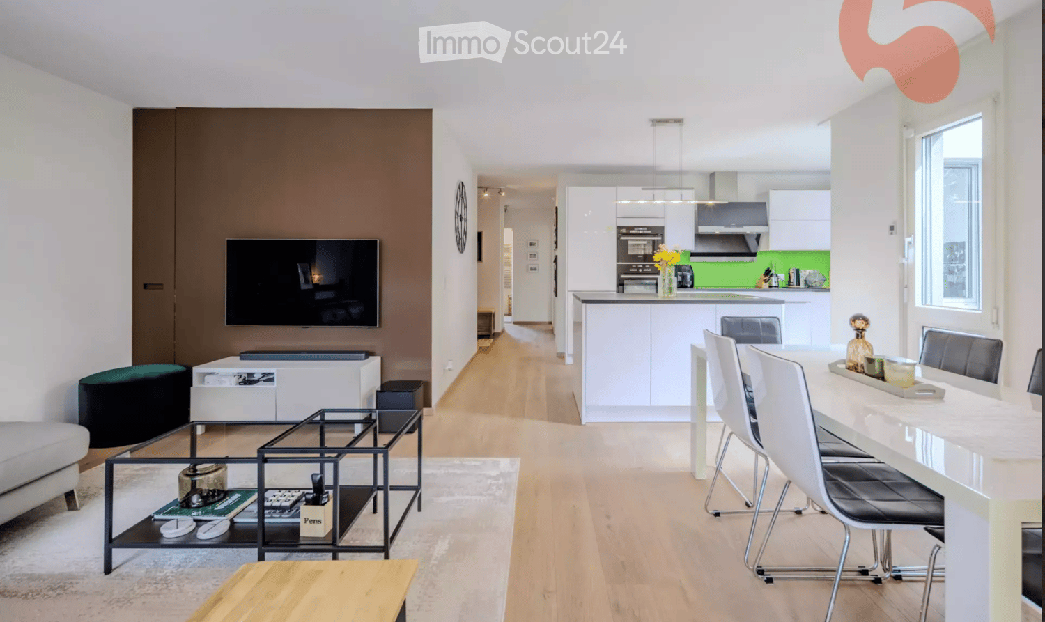 Modern open-plan living and dining area with a sofa, coffee table, TV, and a dining table with chairs. The kitchen features white cabinets, a kitchen island, and various appliances.