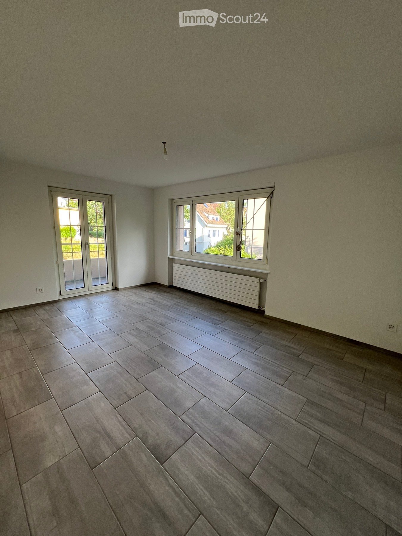 Empty room with light grey tiled flooring, white walls, and large windows with white frames. A radiator is visible beneath one window, and a ceiling light fixture is present.