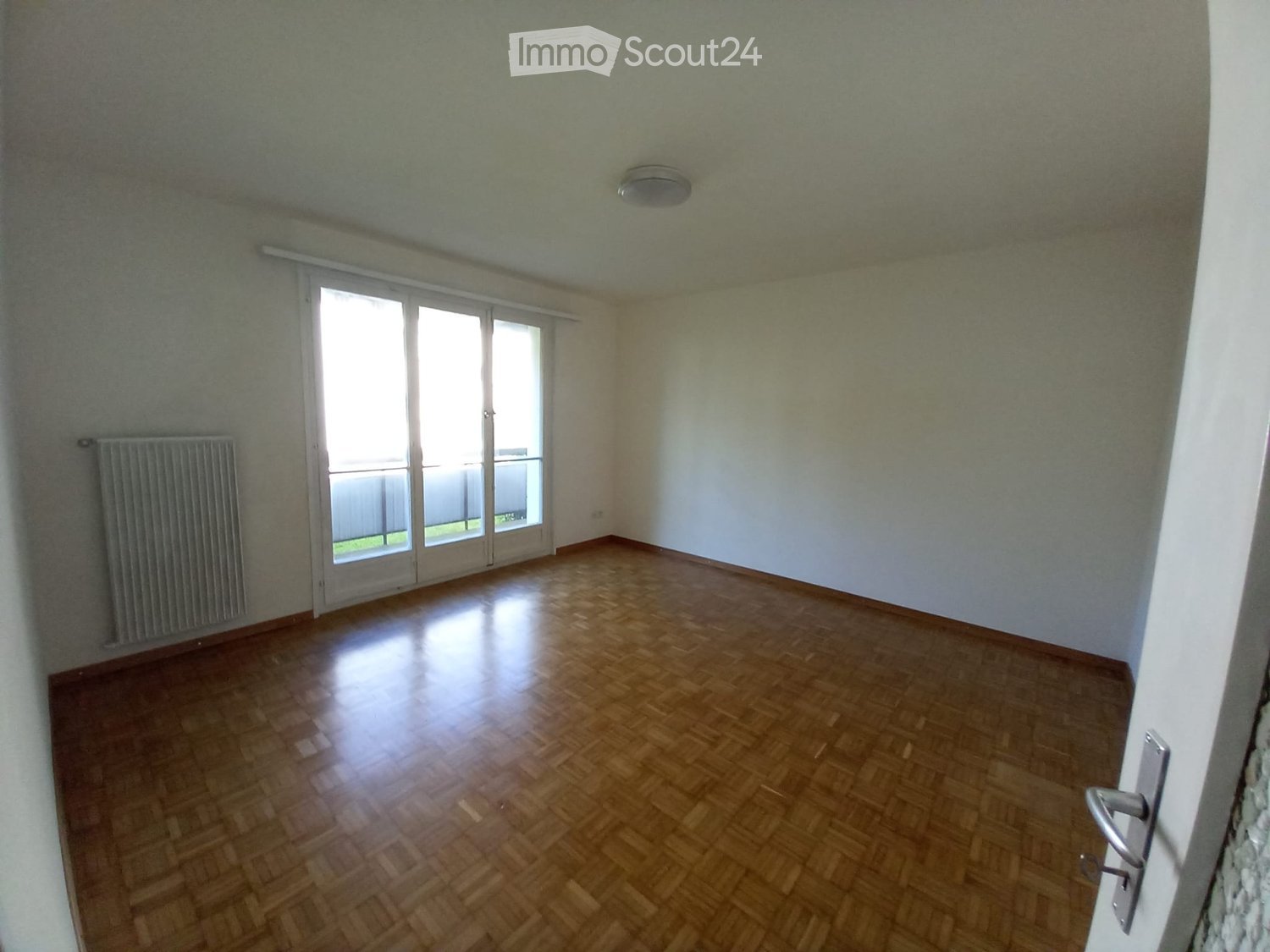 Empty room with wooden flooring, white walls, large sliding glass door leading to a balcony, and a radiator on one wall.