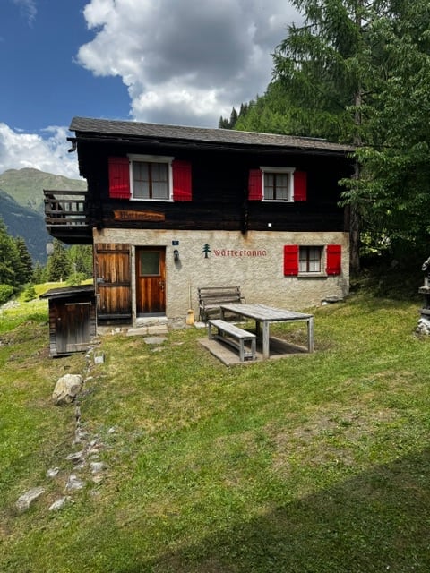 Traditional wooden chalet with red shutters, stone walls, and a balcony overlooking a mountain view. Outdoor seating area with a picnic table and benches.
