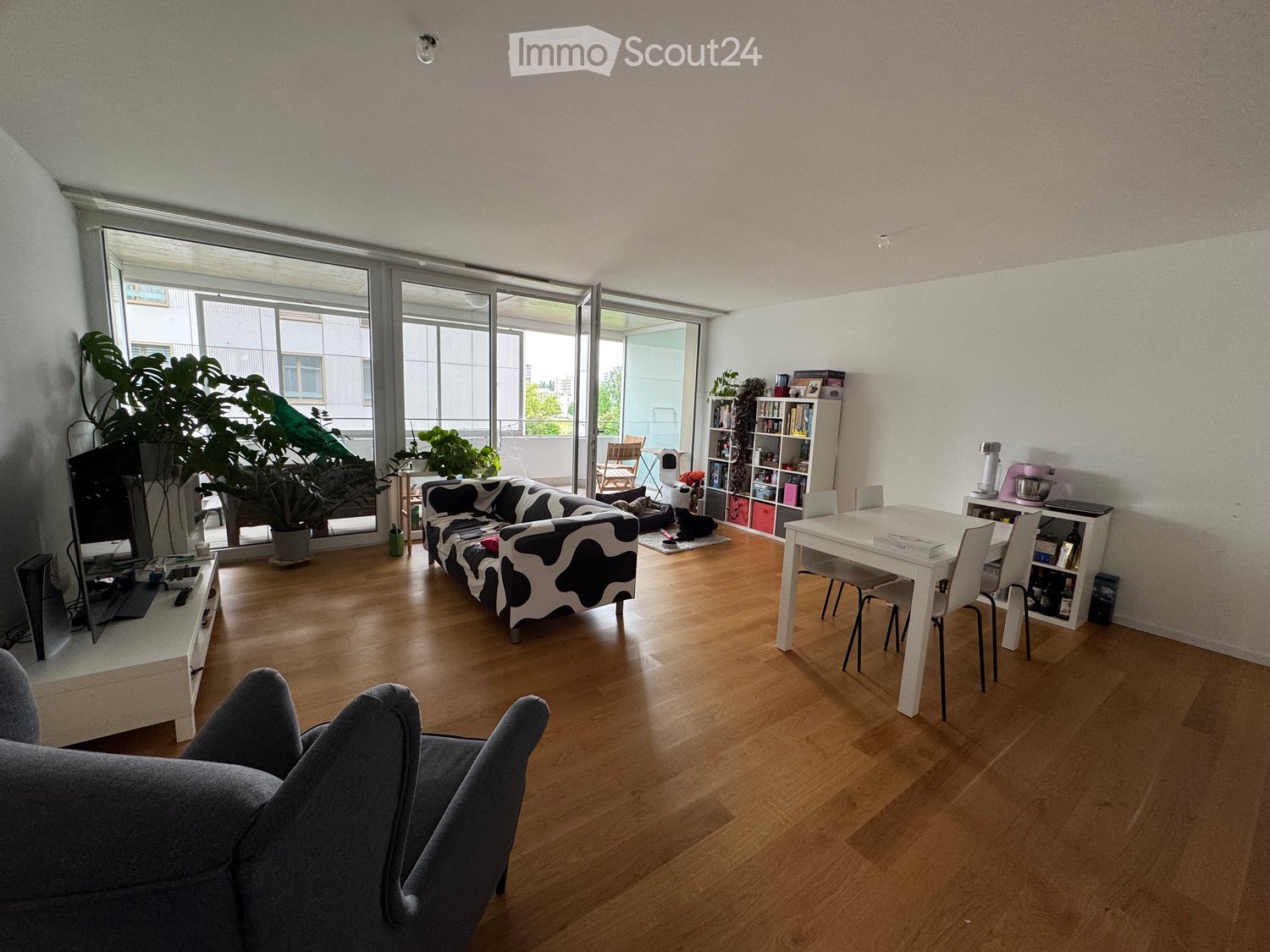 Bright living room with wooden flooring, large windows, and a balcony. Features a black and white cow-print sofa, a grey armchair, a white dining table with chairs, a TV stand, and a bookshelf.