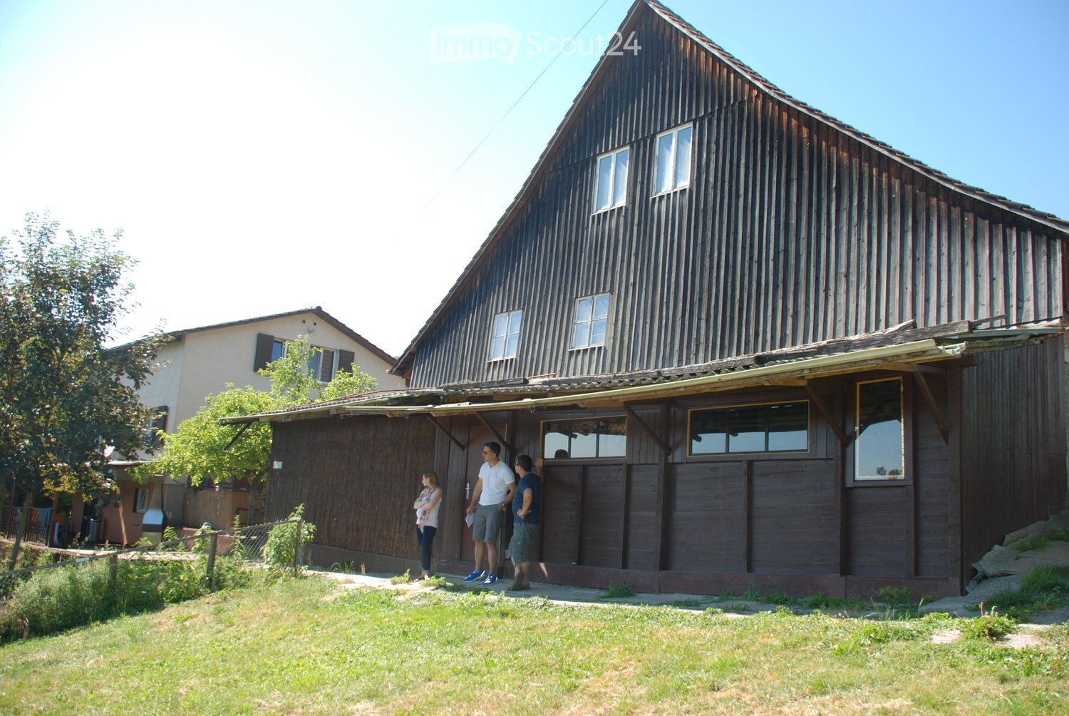 A two-story wooden house with a steep, dark wooden roof and multiple windows. The house has a covered entrance area with large windows and a door. The surrounding area includes a grassy lawn and some trees.
