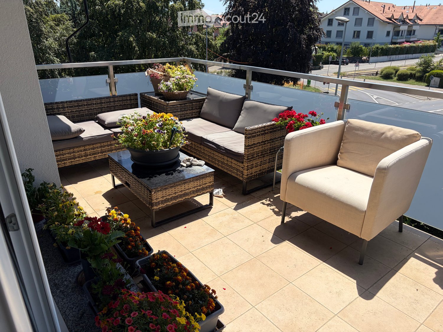 Balcony with a sofa, chair, table, and several flower pots arranged on a tiled floor.