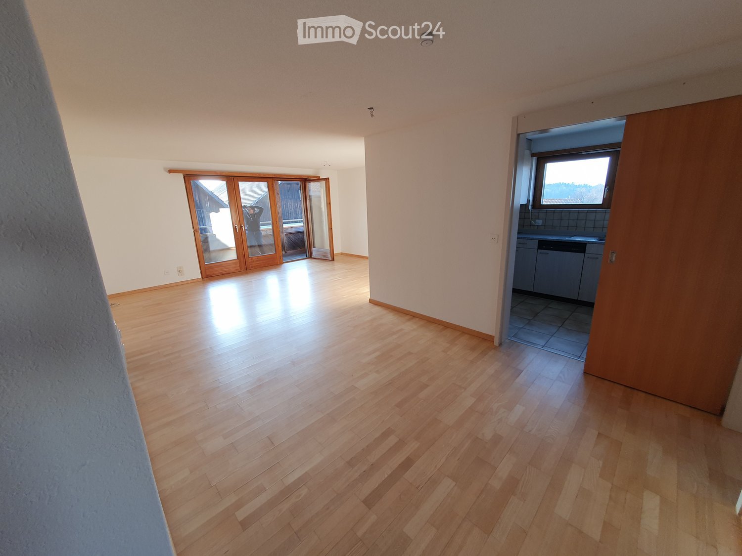 Empty room with wooden flooring, large sliding glass doors leading outside, and an adjacent kitchen area visible through an open doorway.