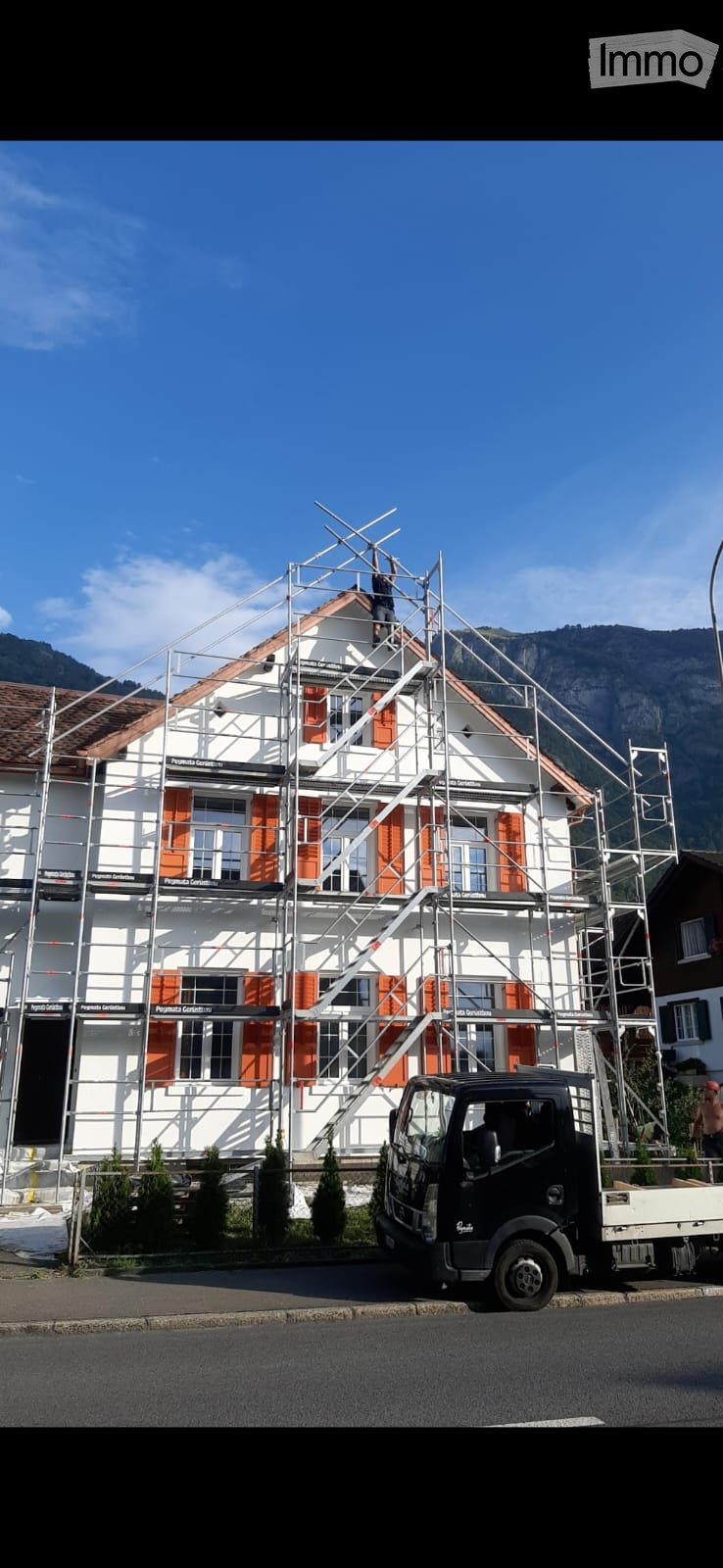 A two-story house under construction with scaffolding, orange shutters, and a worker on the roof. A truck is parked nearby.