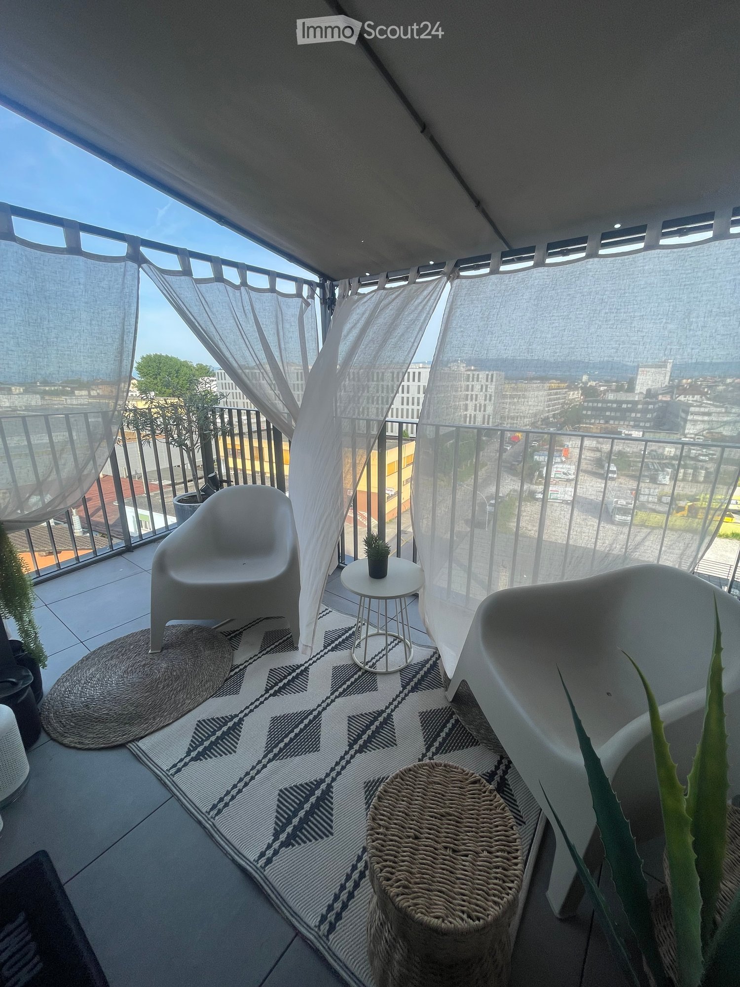 Modern balcony with white furniture, a round table, a woven stool, a patterned rug, and potted plants, offering a city view.
