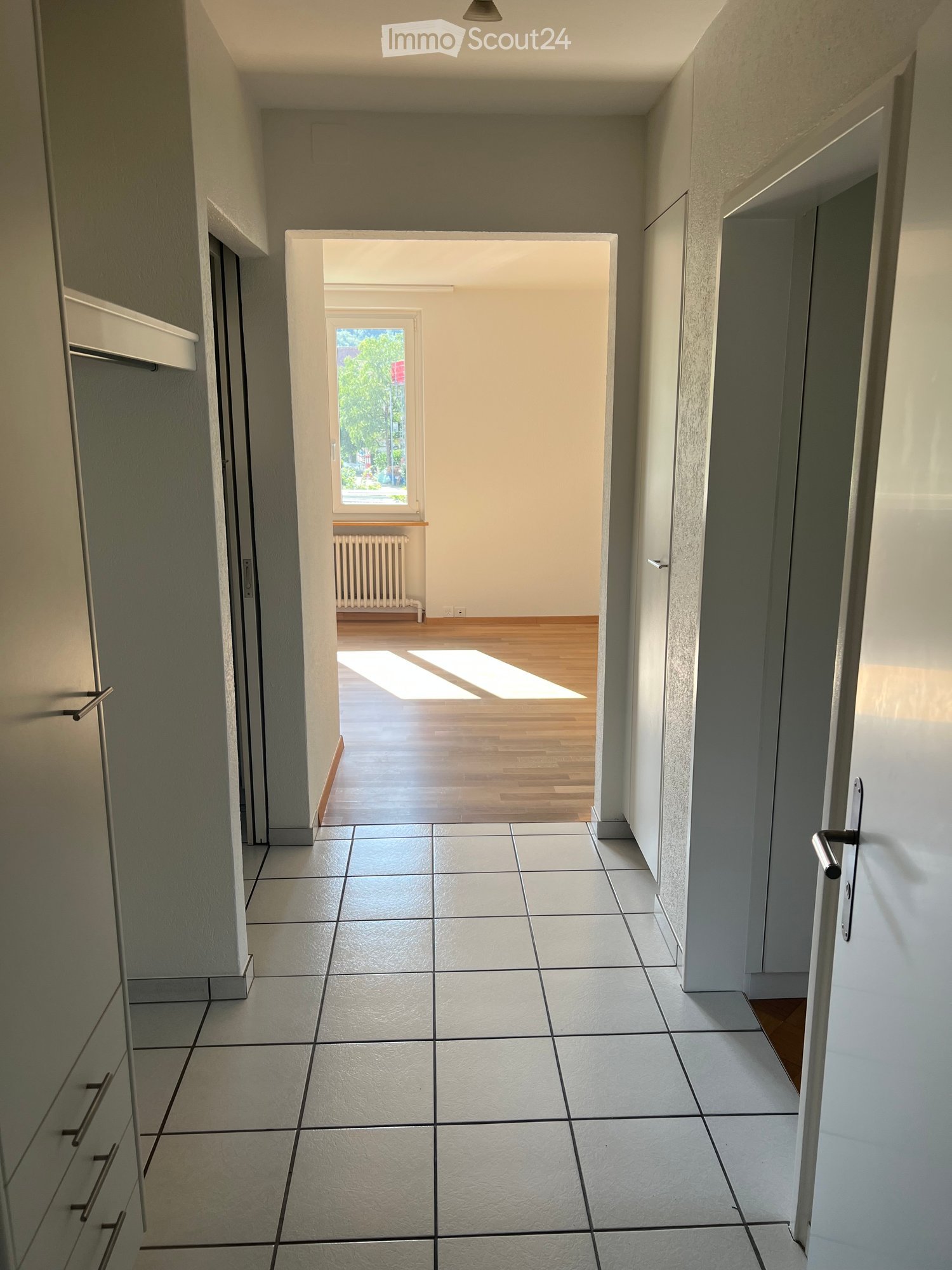 Empty hallway with tiled floor, white walls, and doors leading to other rooms. Natural light streams in from a window at the end.