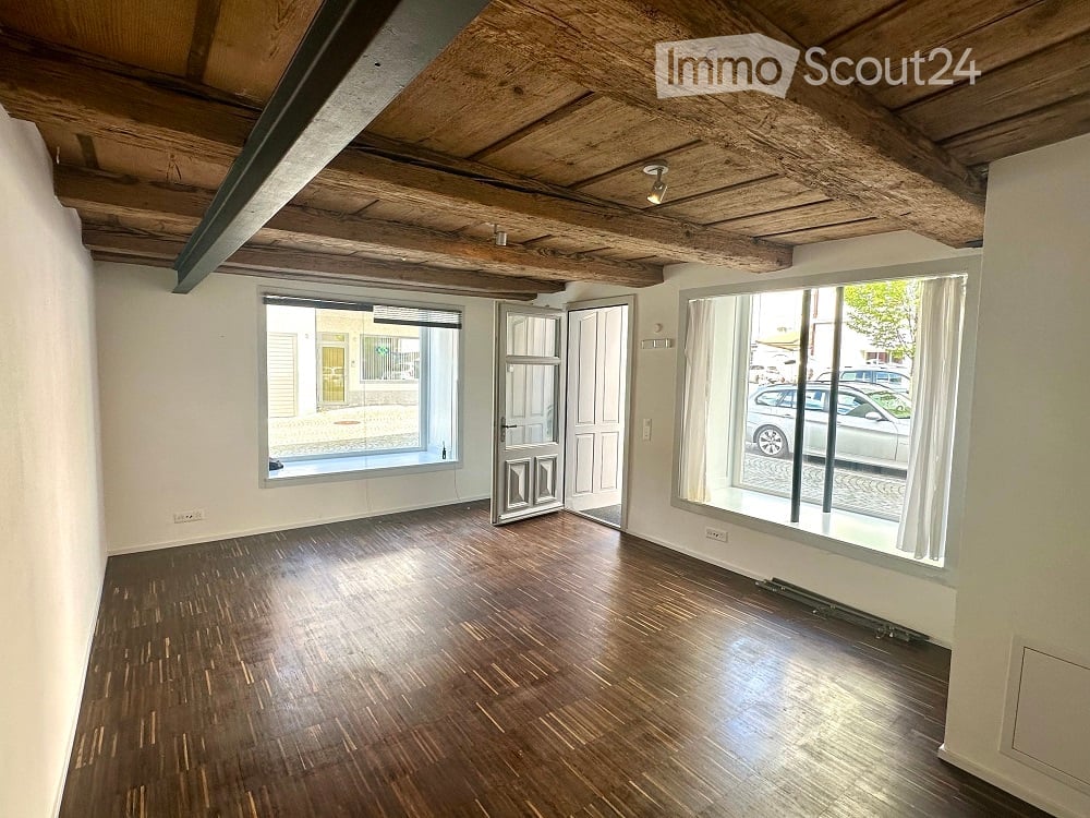 Empty room with wooden flooring, exposed wooden ceiling beams, large windows, and a door leading outside.