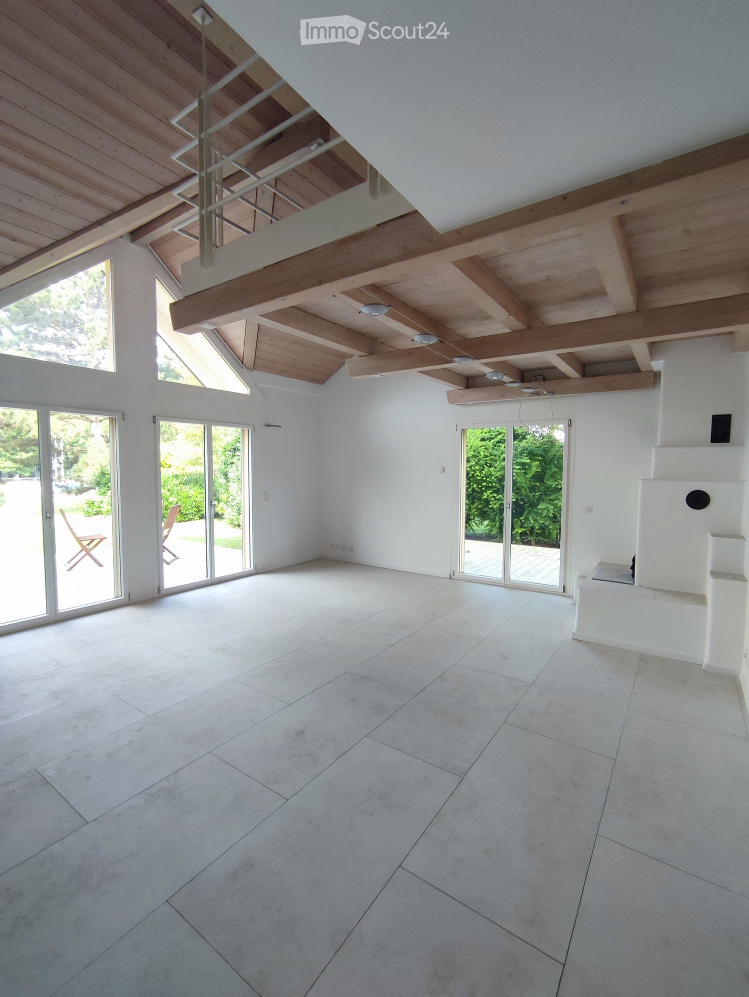 Empty room with white walls, large skylights, and wooden beams on the ceiling. The floor is tiled, and there are large glass doors leading to an outdoor area.