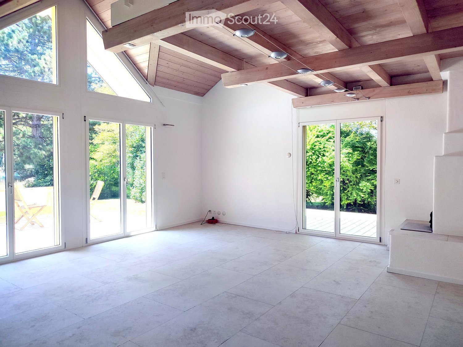 Bright, open room with white walls, exposed wooden ceiling beams, large glass doors leading to outdoor space, and a tiled floor.