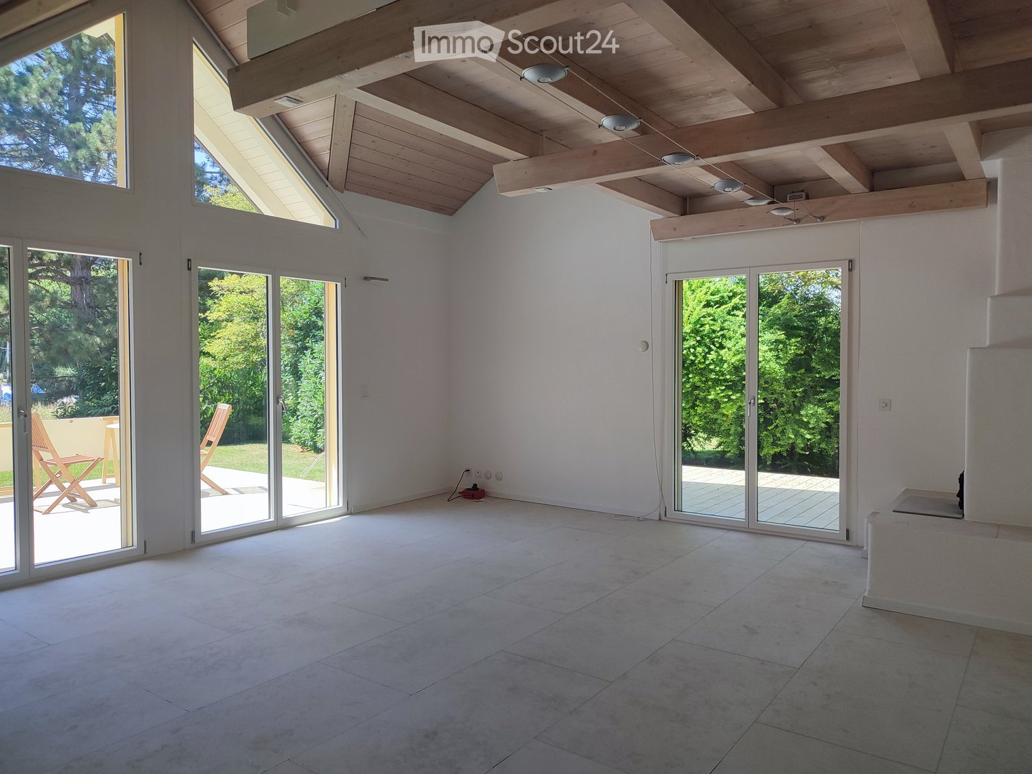 Bright, open room with large glass doors leading to an outdoor area, exposed wooden ceiling beams, and a smooth tiled floor.