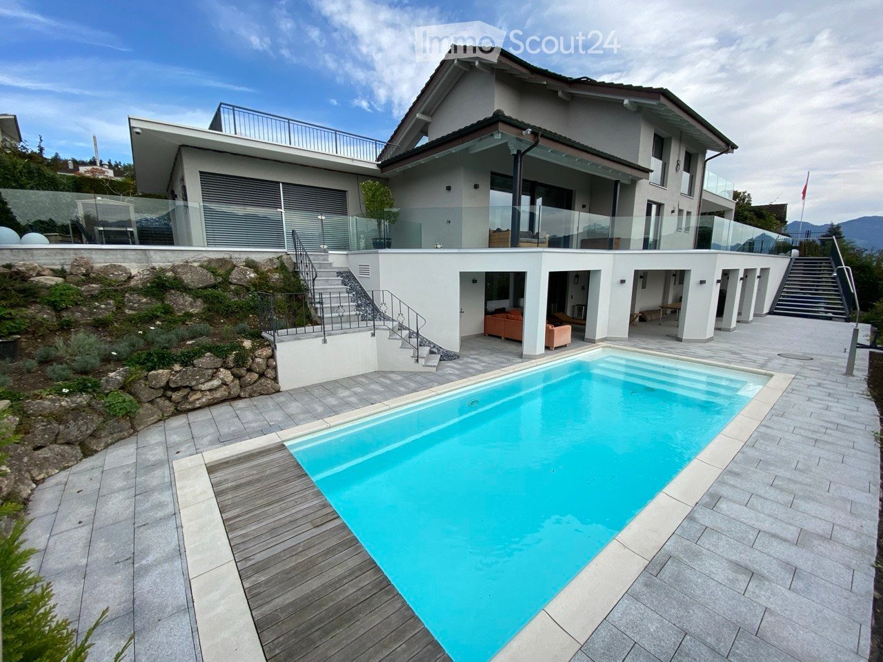 Modern house with a swimming pool in the foreground, surrounded by a paved patio and steps leading up to the house. The house features large glass windows and a balcony on the upper level.