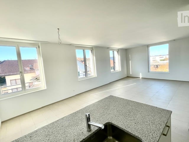 Empty room with white walls, tiled floor, and large windows. A kitchen counter with a sink and faucet is visible.