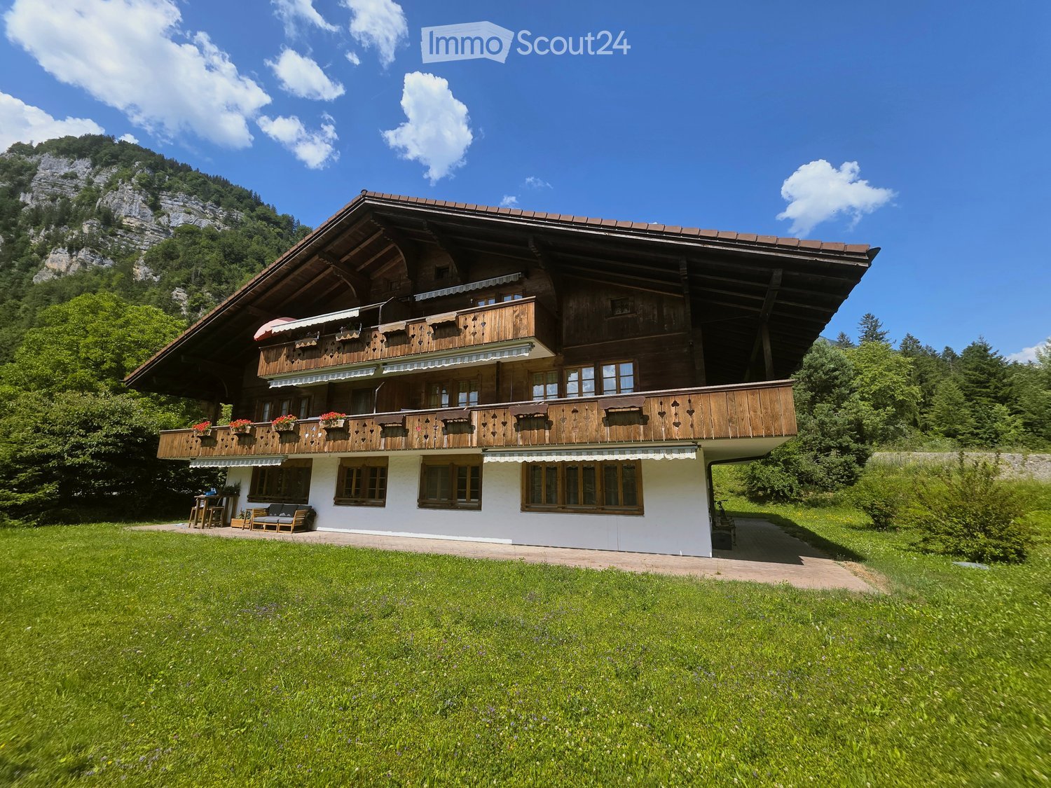 A large, two-story wooden house with balconies, surrounded by a green lawn and mountains in the background.