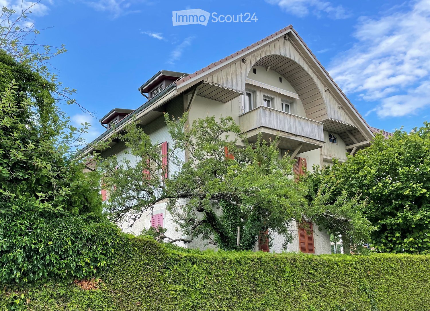 Two-story house with white walls, red shutters, and a balcony on the upper floor, surrounded by greenery and a hedge.