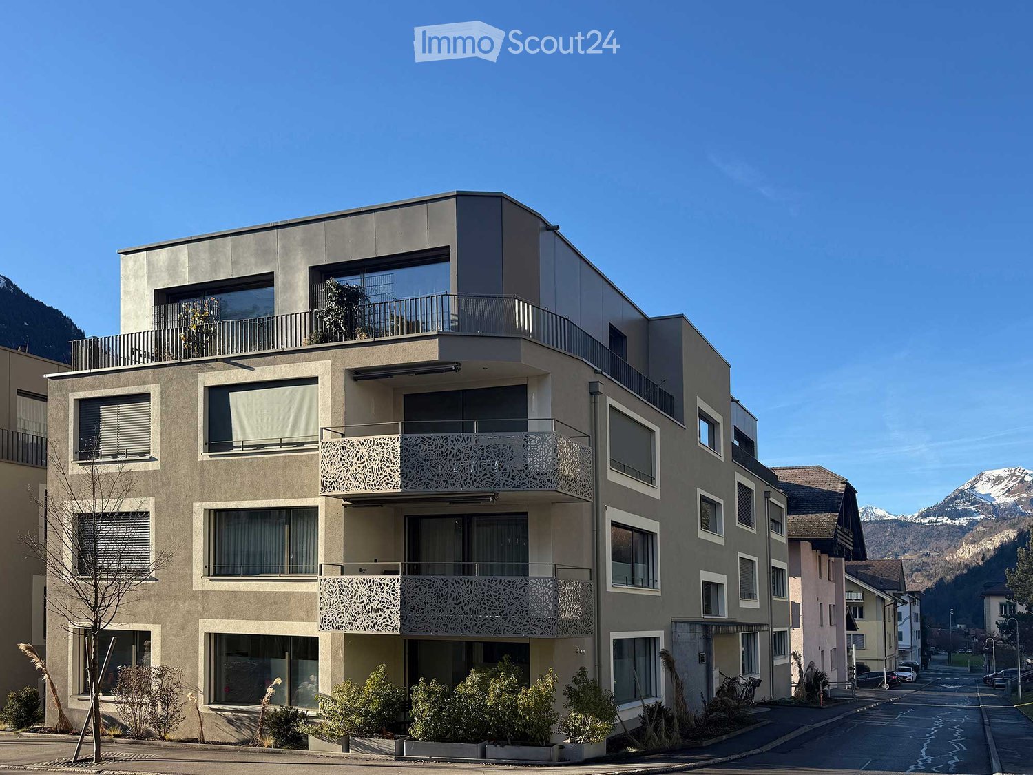 4-story building with multiple balconies, grey facade, glass windows, and green landscape