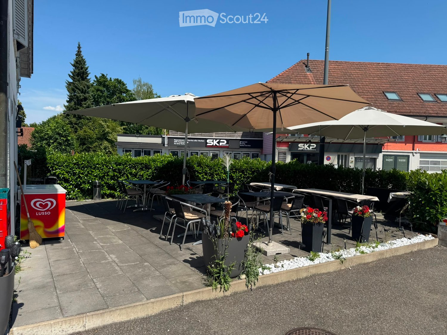 Outdoor seating area with tables, chairs, and umbrellas, surrounded by greenery and a building in the background.