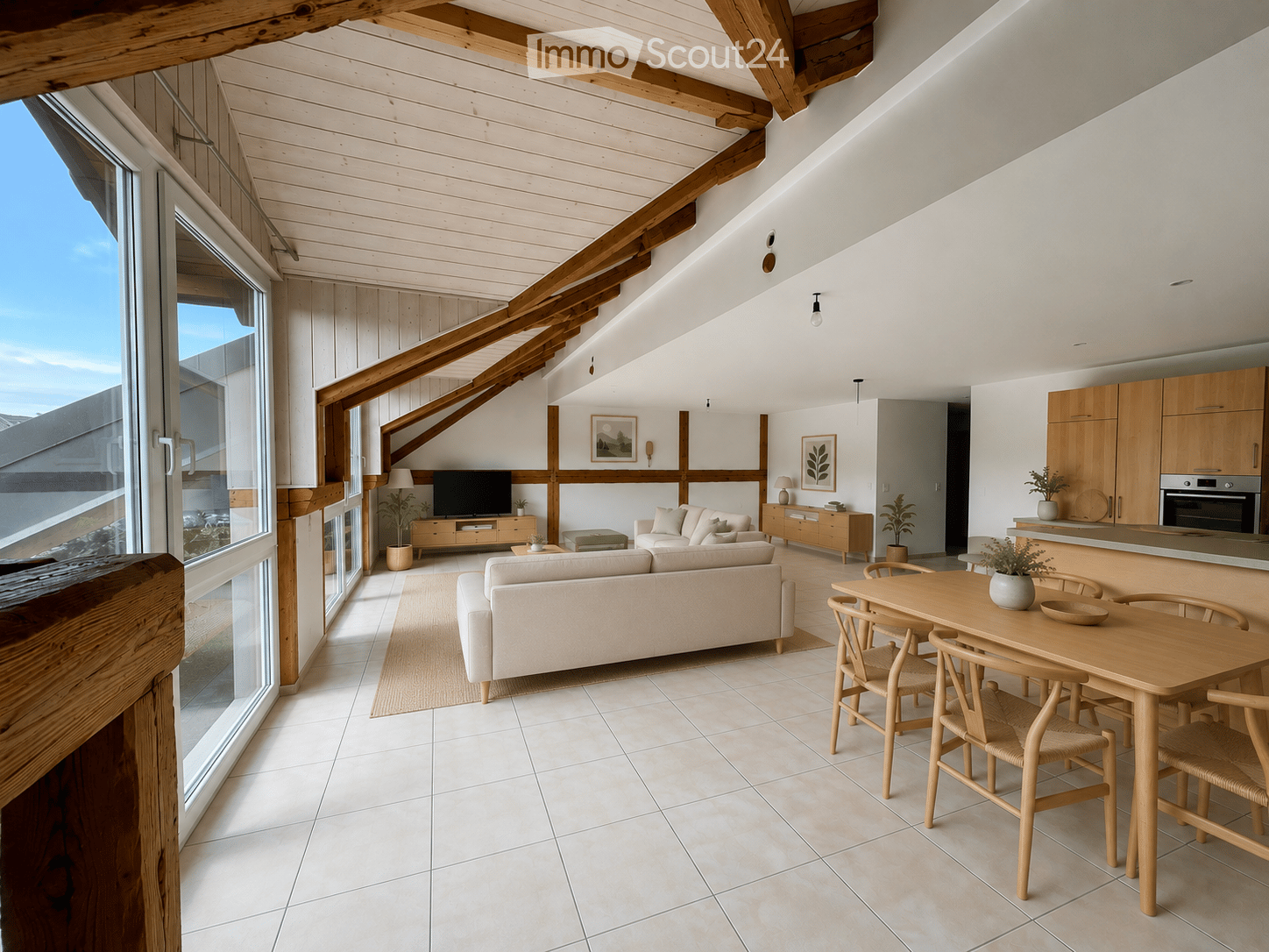 Bright, airy living space with large windows offering a scenic view, featuring a cream-colored sofa, wooden dining table with chairs, a television, and modern kitchen cabinets.