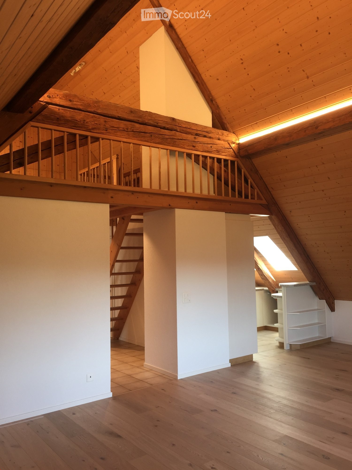 An attic space with wooden beams, exposed wooden ceiling, wooden floor, and a staircase leading to an upper level. The room is empty with white walls and a small window letting in natural light.