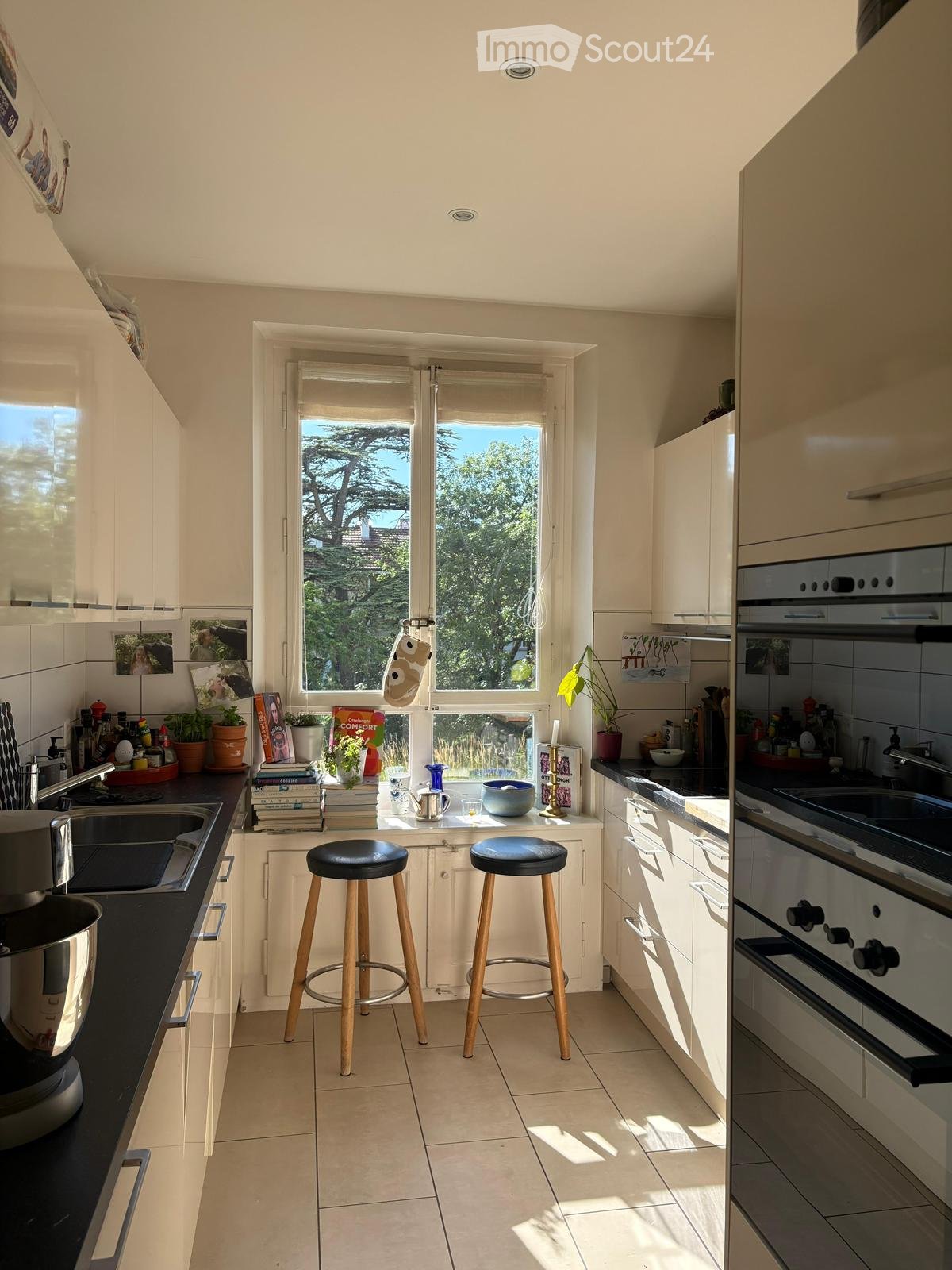 Bright kitchen with white cabinets, black countertops, and a large window overlooking greenery. Two wooden stools are placed in front of the island, and various kitchen items, including a sink, oven, and potted plants, are visible.