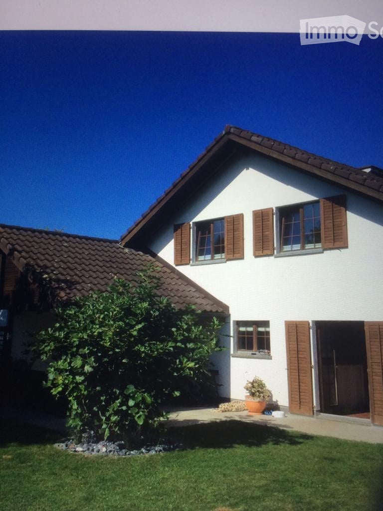 Two-story white house with brown shutters and tiled roof, surrounded by a well-maintained lawn and small garden area.