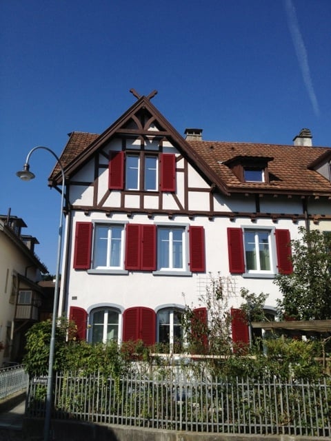 Two-story house with white walls, red shutters, and a brown tiled roof. The house features a decorative wooden gable and is surrounded by a metal fence and greenery.