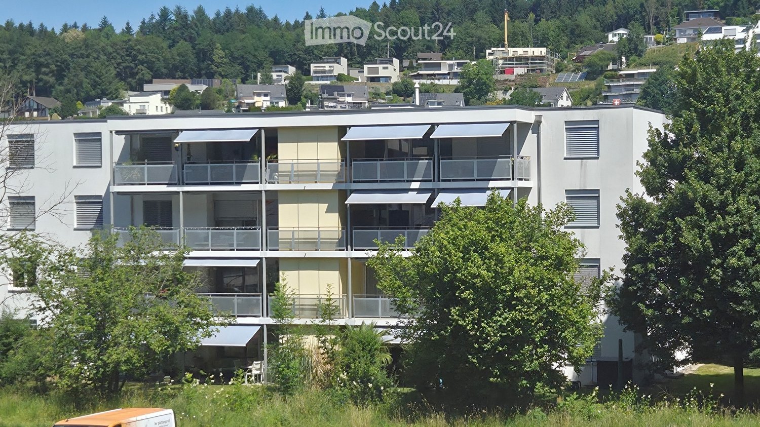Modern multi-story residential building with balconies, awnings, and surrounding greenery.