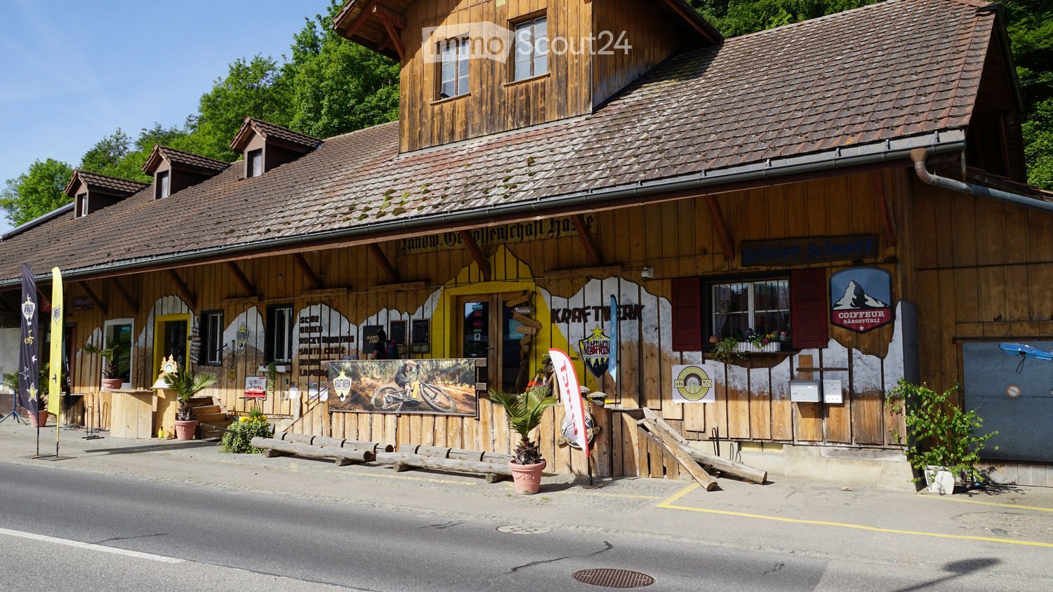 A rustic wooden building with a tiled roof, signage for "Kraftwerk" and "Coiffeur Mangstuli", outdoor seating area with wooden benches, potted plants, and banners.