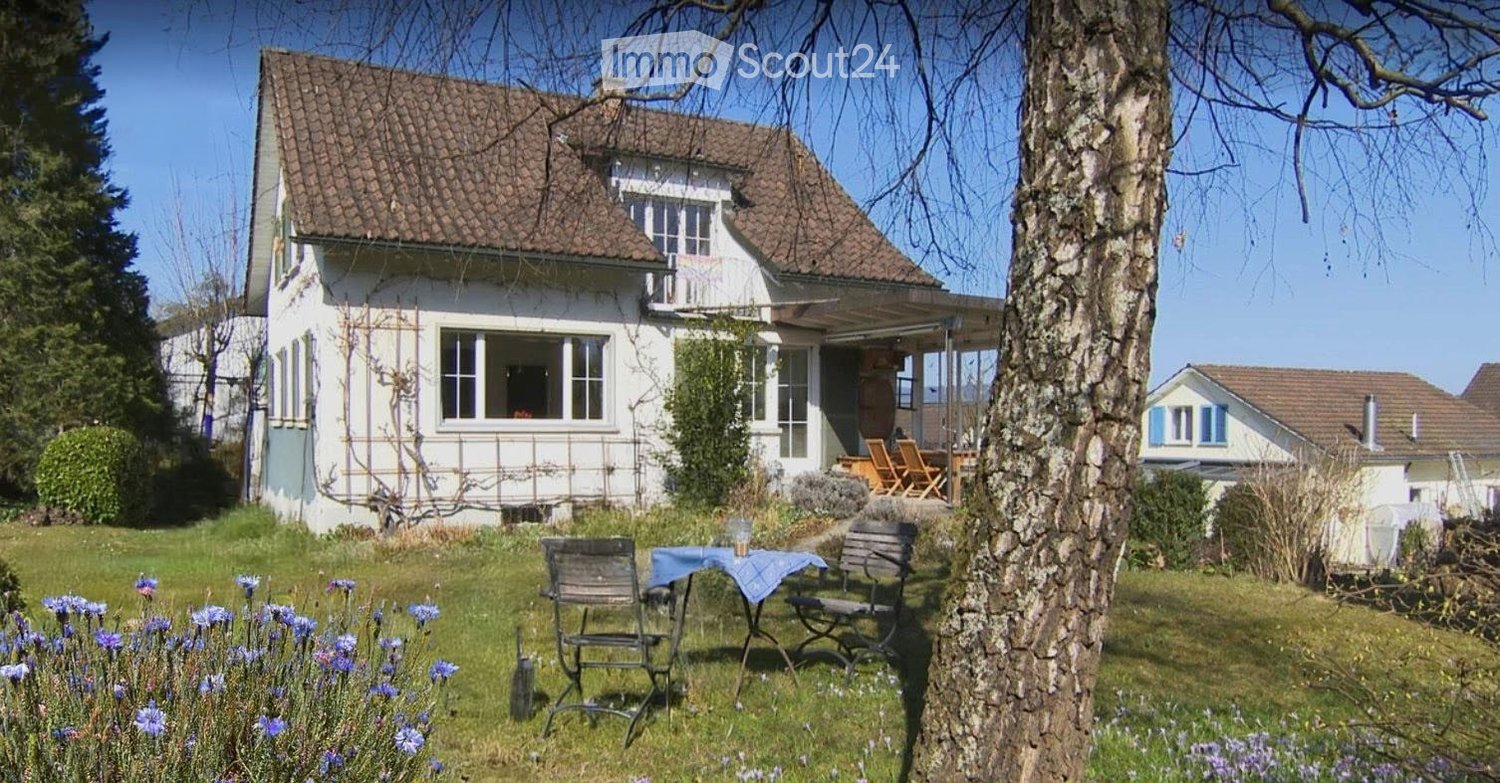 Garden with a table and chairs, surrounded by flowers and trees, with a house in the background.