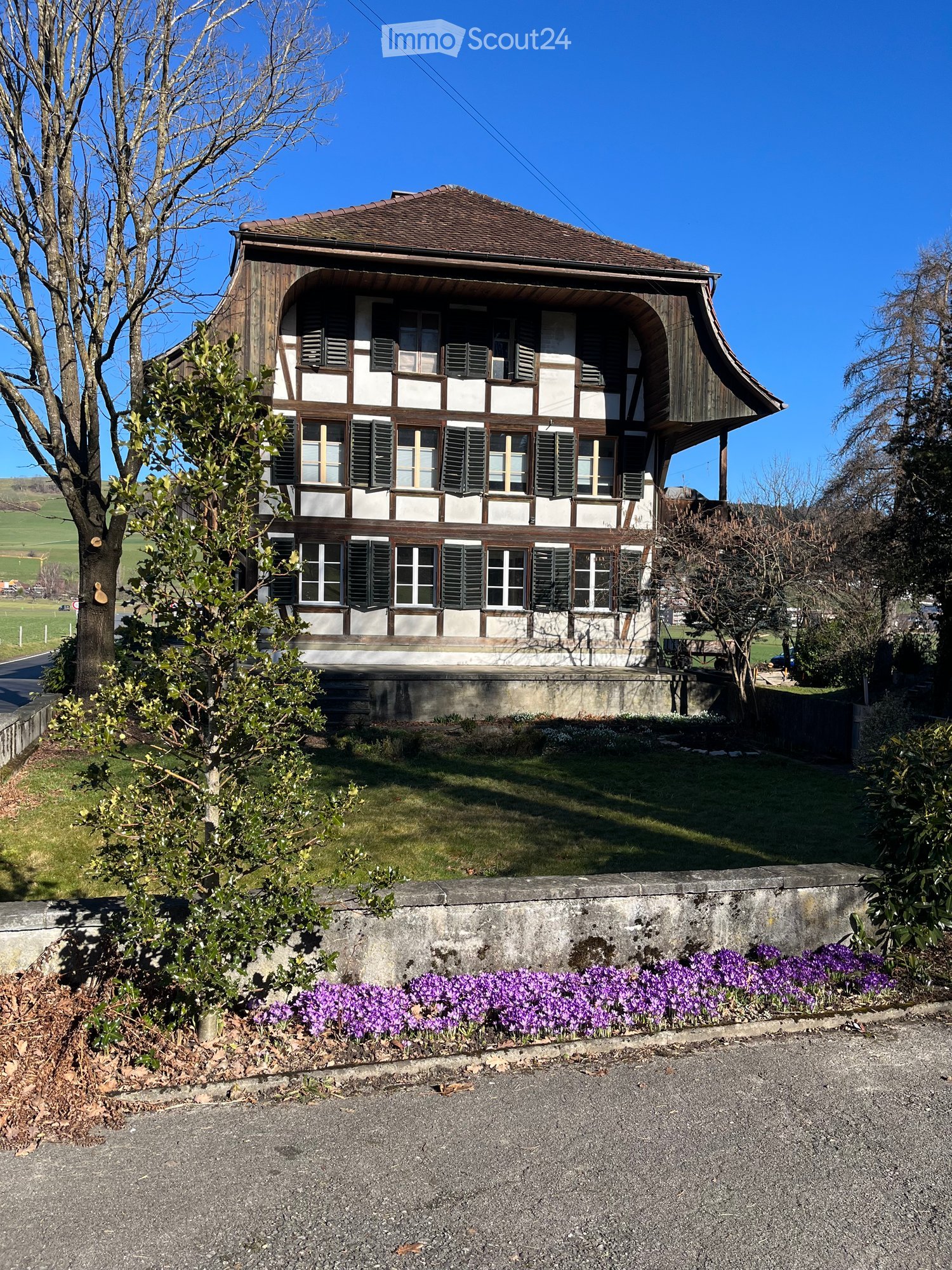 Two-story traditional house with wooden beams, white walls, multiple windows, and a raised ground floor. Surrounded by greenery and purple flowers.