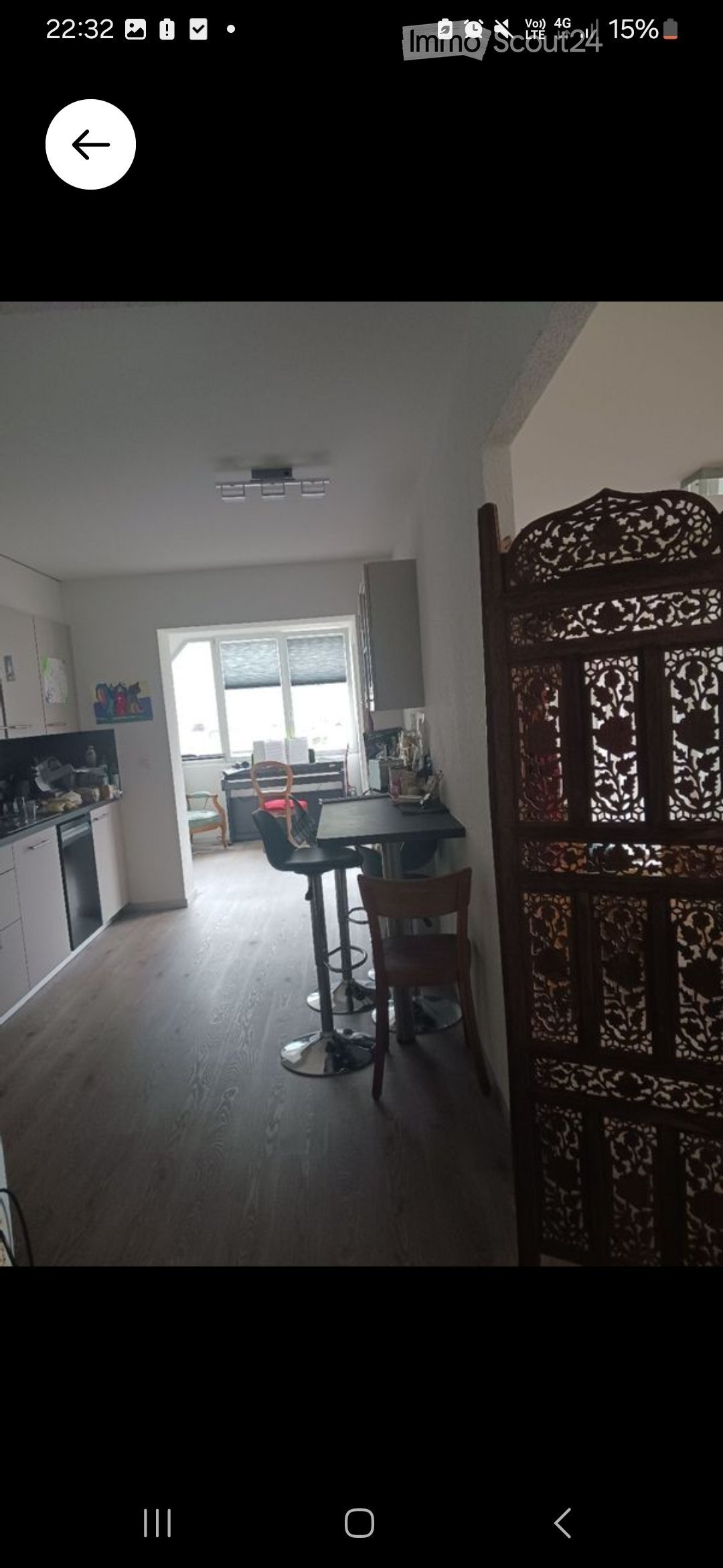 A modern, open-plan living and dining area with wooden flooring, white kitchen cabinets, a dining table with chairs, and a decorative screen. The space is well-lit with natural light from large windows.