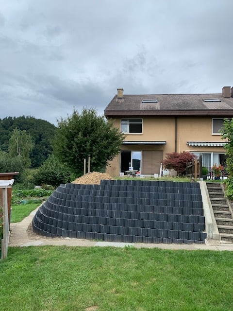 A house with a well-maintained garden, featuring a curved retaining wall made of dark bricks, a staircase leading up to the house, and a small patio area with outdoor furniture.
