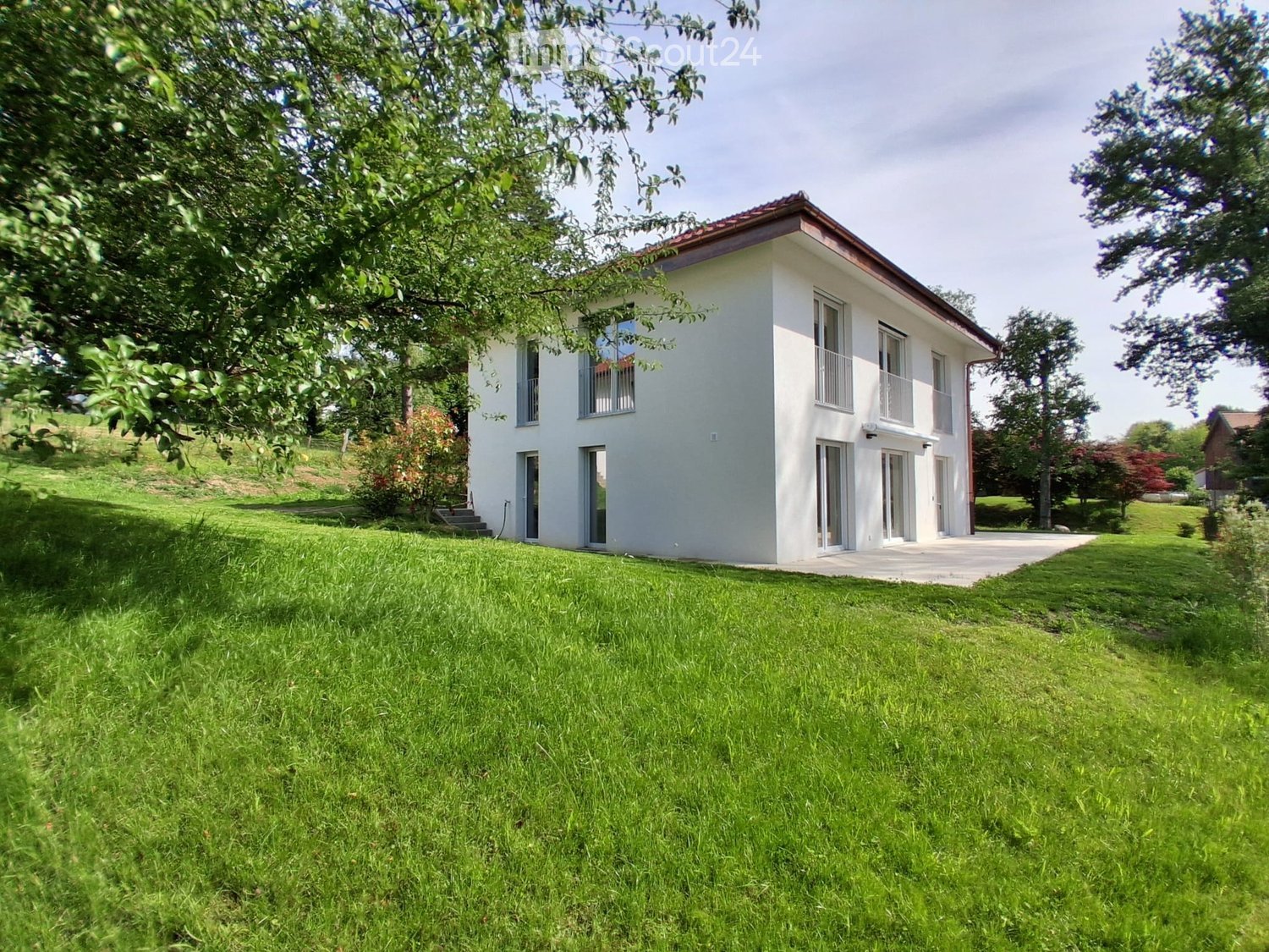 Modern two-story house with white walls, large windows, and a red-tiled roof, surrounded by a well-maintained lawn and trees.