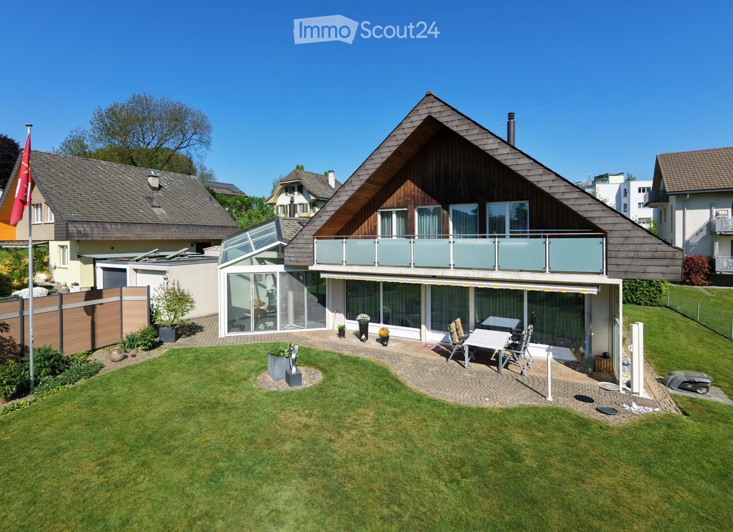 A modern two-story house with a triangular roof, large windows, and a spacious balcony. The property features a well-maintained lawn, patio area with outdoor furniture, and surrounding greenery.