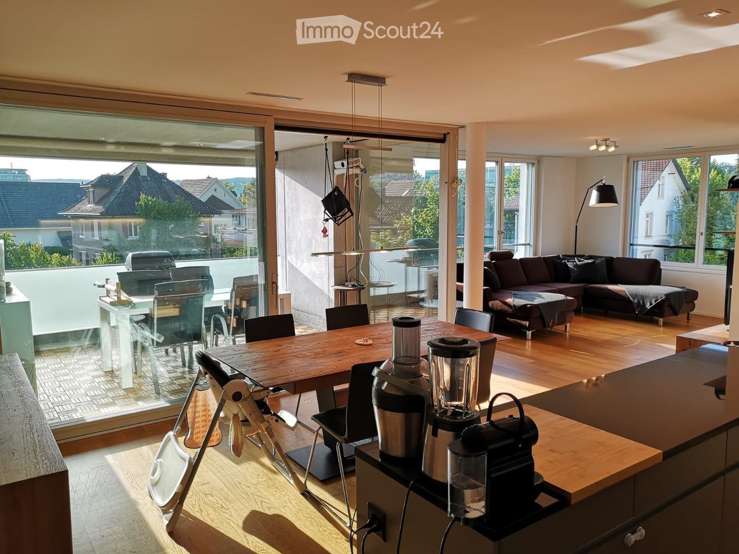 Open-plan living and dining area with wooden flooring, large windows, a sofa, a coffee table, a dining table with chairs, and a kitchen counter with a coffee machine and blender.
