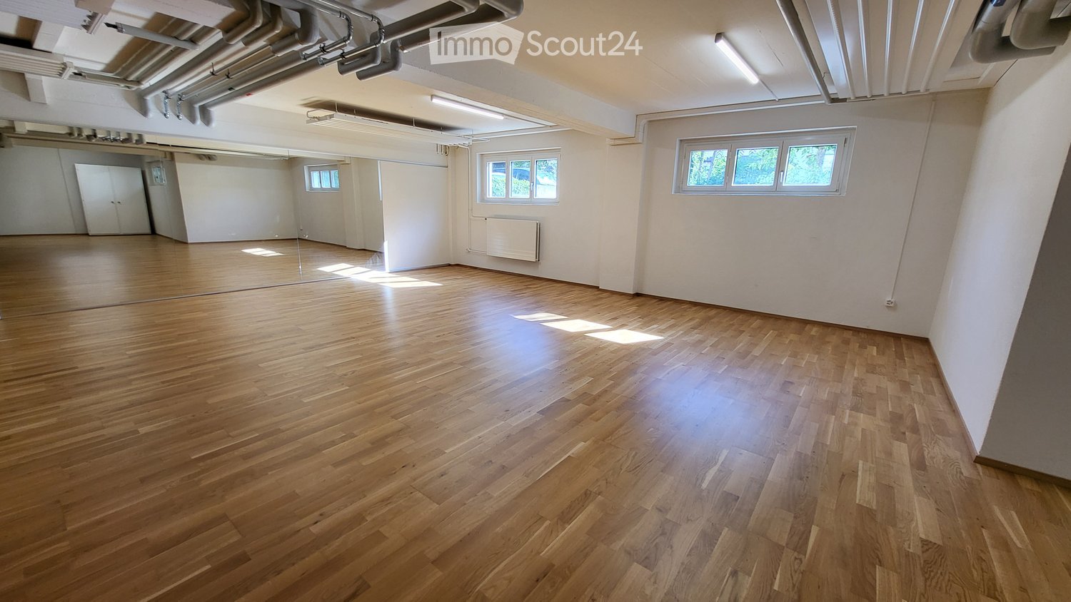 Empty room with wooden flooring, white walls, and large windows allowing natural light. No furniture or equipment present.